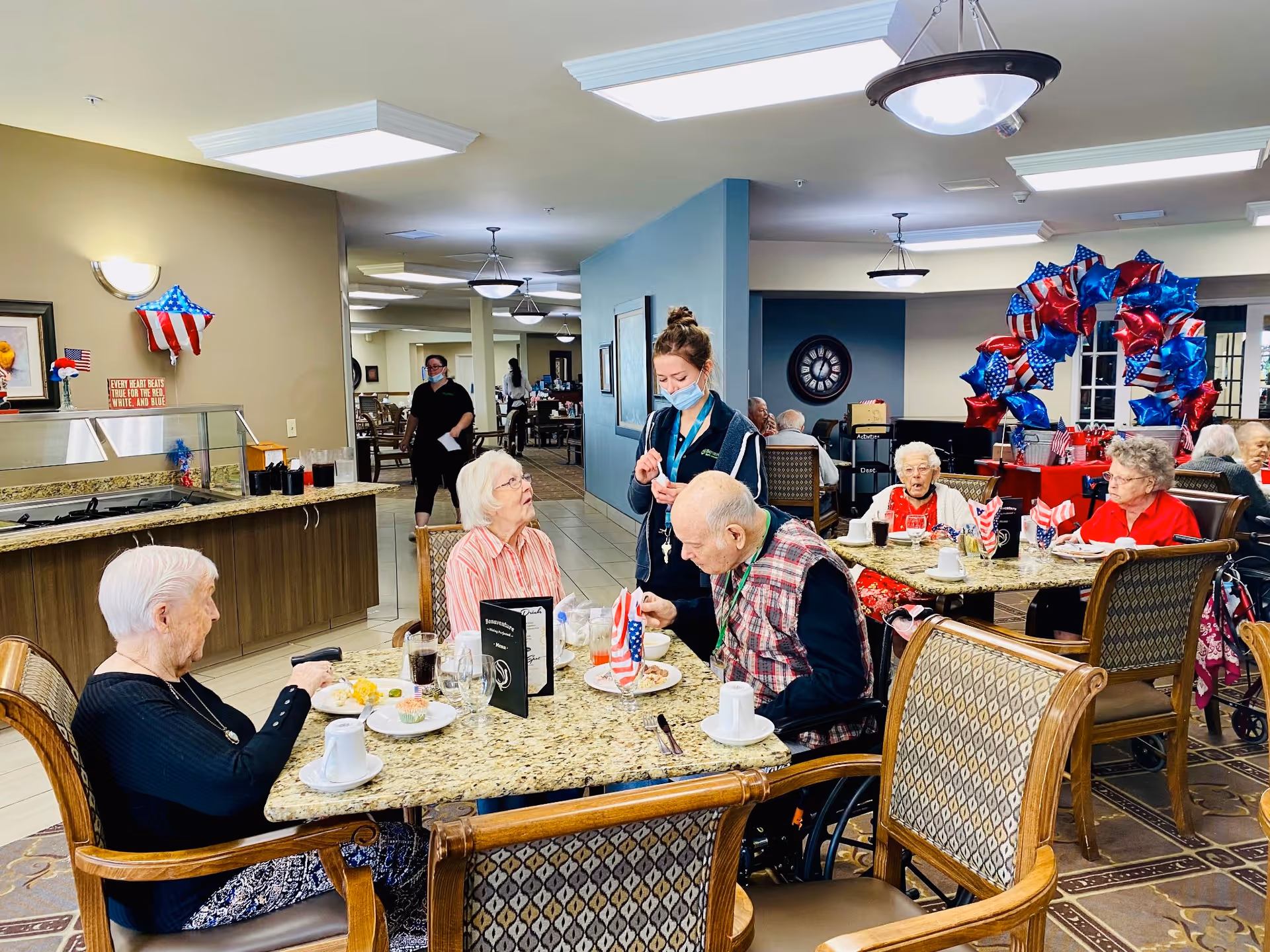 A dining room in a senior living facility where elderly residents are seated at tables enjoying a meal. A staff member wearing a mask is interacting with one of the residents. The room is decorated with red, white, and blue patriotic balloons and small American flags on the tables.