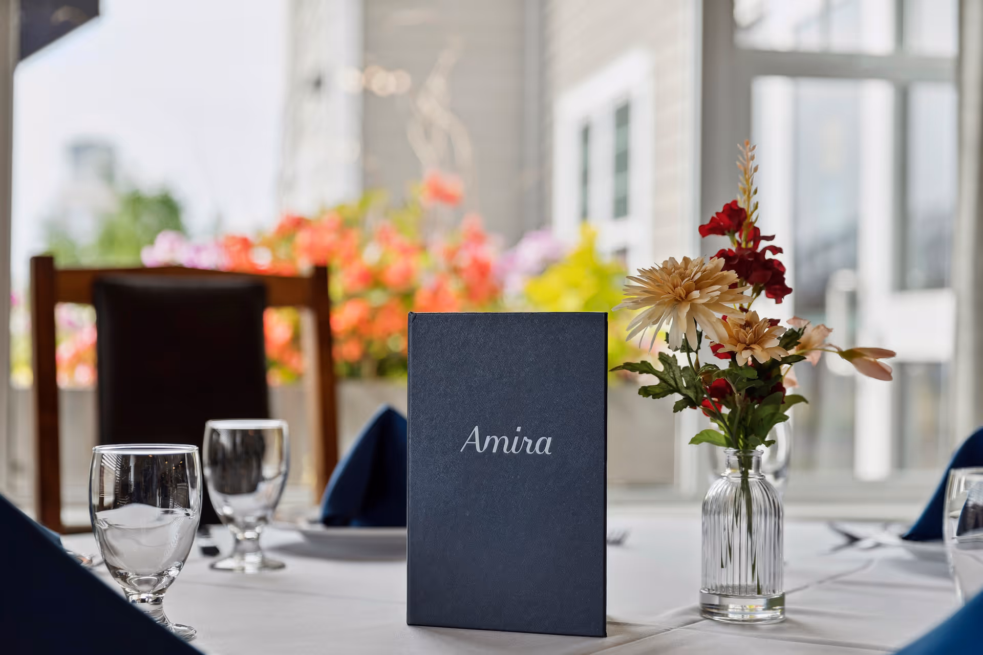 A dining table set with clear water glasses, navy blue napkins, and a small vase with red and beige flowers. A dark blue menu card with the word 'Amira' is placed in the center of the table. In the background, there are colorful flowers outside a window and a wooden chair.