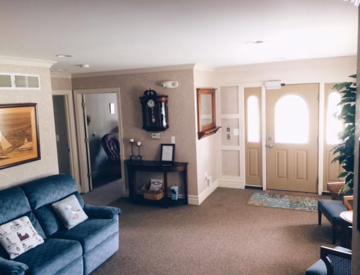 A cozy living room area in Pineview Manor featuring a blue upholstered sofa with decorative pillows, a wooden side table with candles and a framed picture, a wall-mounted clock, a framed painting of a sailboat, and a beige front door with windows letting in natural light. There is also a small bench and a potted plant near the entrance.