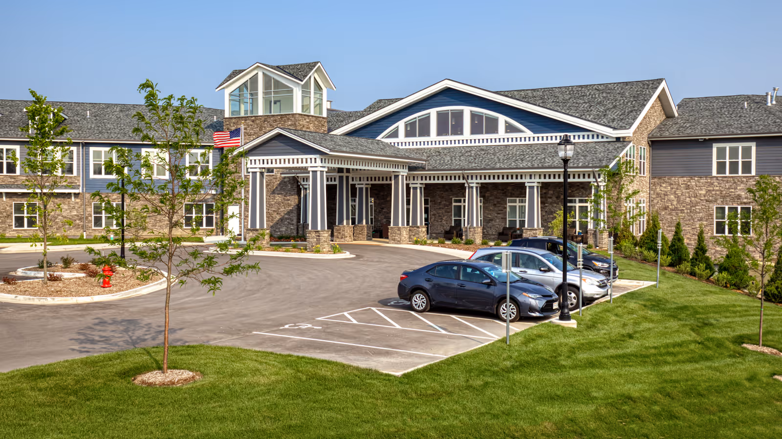 Front exterior of a two-story senior living building with a covered entrance, parked cars, young trees, and an American flag.