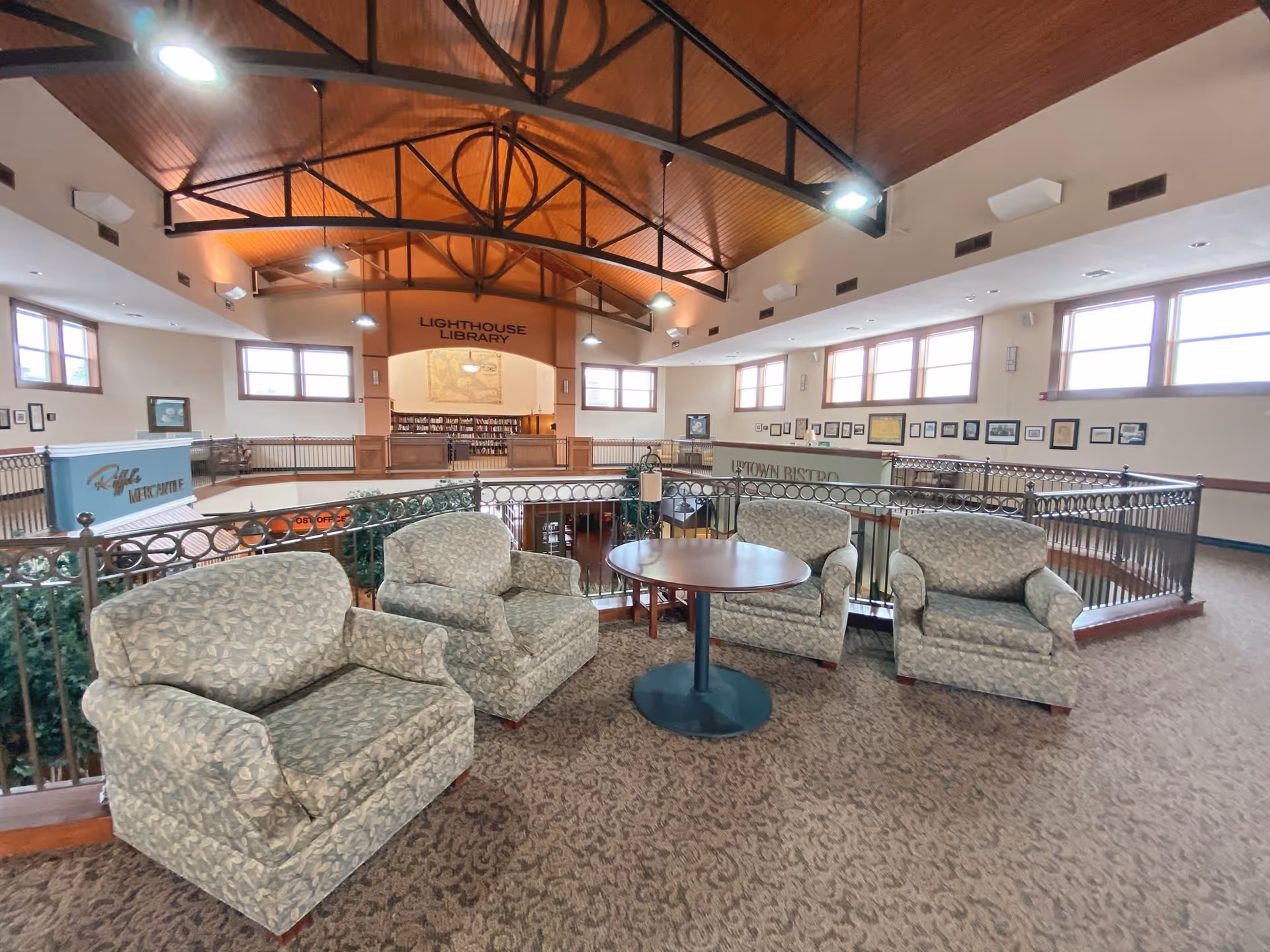 A spacious indoor common area with four patterned armchairs arranged around a round wooden table. The area overlooks a lower level with signs indicating a Lighthouse Library and Uptown Bistro. The ceiling is high with exposed wooden beams and multiple windows allowing natural light to fill the space.
