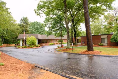 Wet driveway curves toward the entrance of a brick senior living center amid green trees.