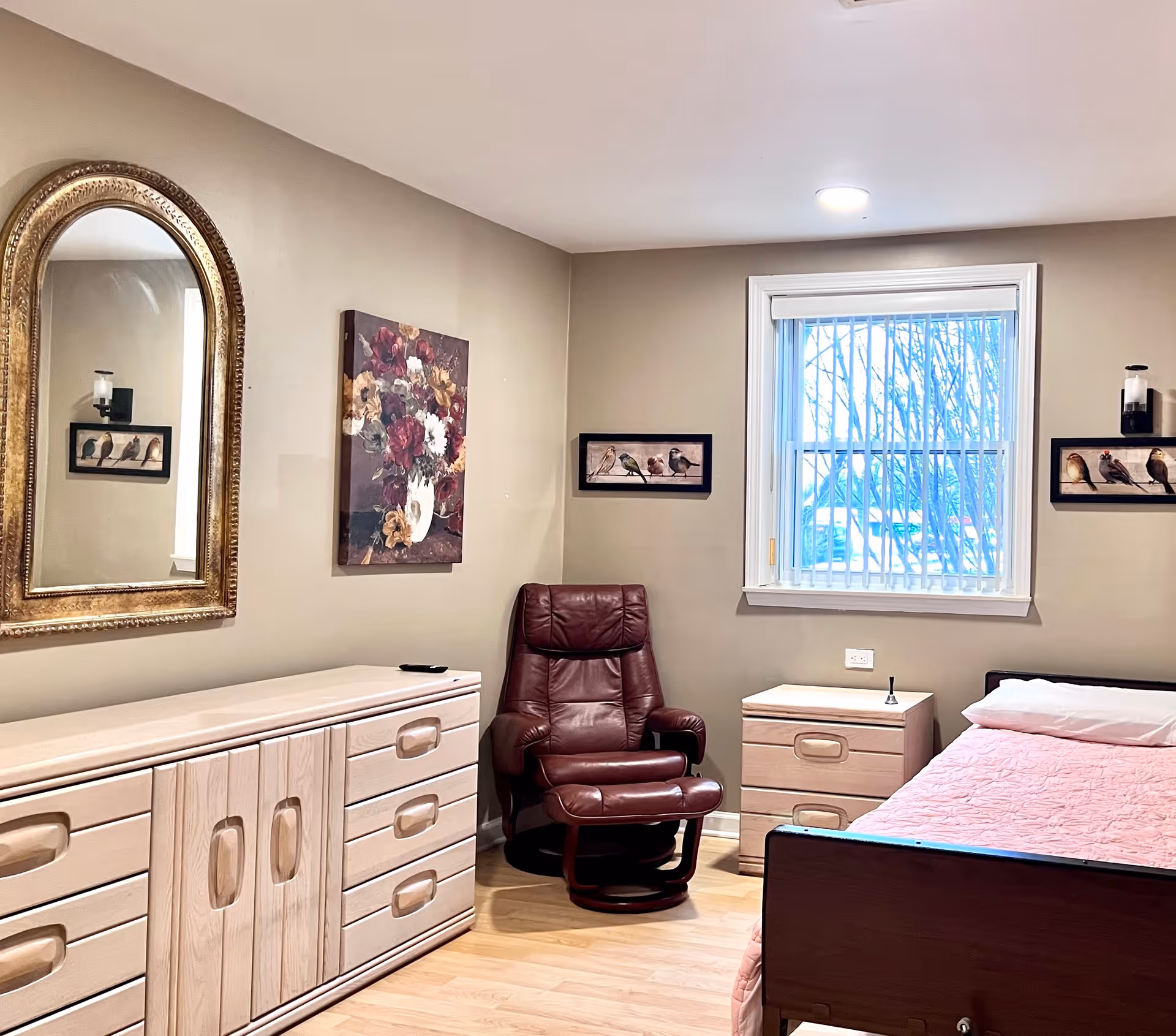 A cozy bedroom in a nursing home featuring a single bed with a pink quilt, a wooden dresser with a large ornate mirror above it, a maroon leather recliner chair, a small wooden nightstand with a bell on top, and a window with vertical blinds. The walls are decorated with framed pictures of birds and a floral painting.