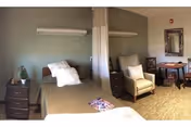 A shared bedroom in a senior living facility with two beds separated by a privacy curtain. Each bed has a bedside table with a lamp and personal items. There is a comfortable armchair and a small table with chairs near a wall with a mirror.