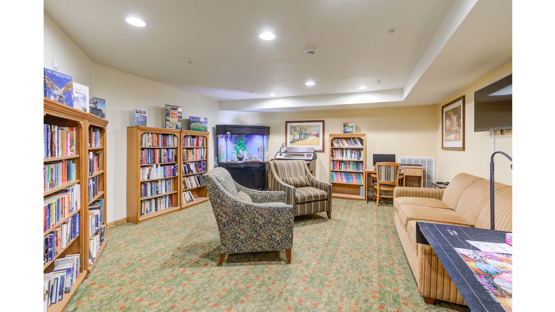 A cozy reading and relaxation room with multiple wooden bookshelves filled with books, two patterned armchairs, a striped sofa, a fish tank, a small desk with a chair, and framed artwork on the walls. The room has a floral carpet and recessed ceiling lights.