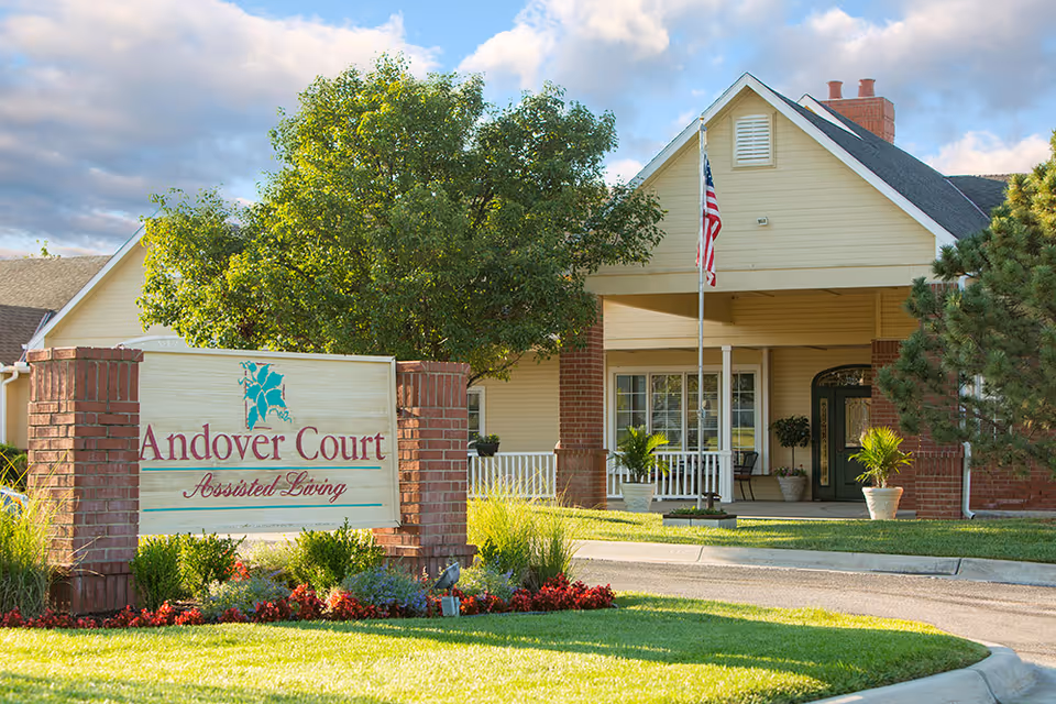 Exterior view of Andover Court Assisted Living facility with a large sign in front surrounded by flowers and greenery. The building has a covered entrance with brick pillars, potted plants, and an American flag on a flagpole. The sky is partly cloudy.