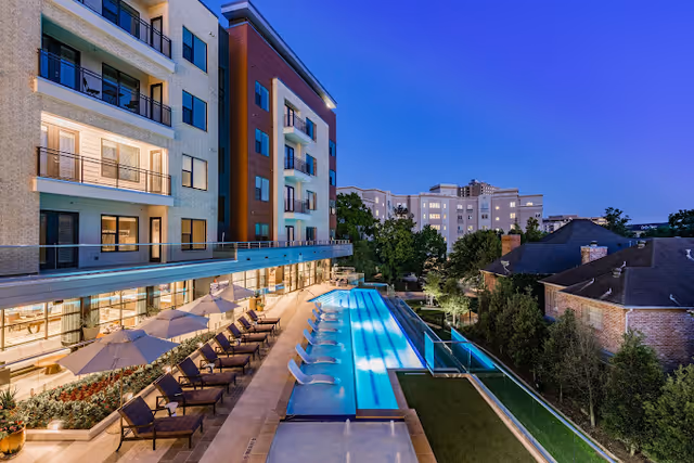 Outdoor view of a modern senior living facility at dusk featuring a long swimming pool with lounge chairs and umbrellas along the poolside. The building has multiple floors with balconies and large windows, and there are trees and neighboring buildings in the background.
