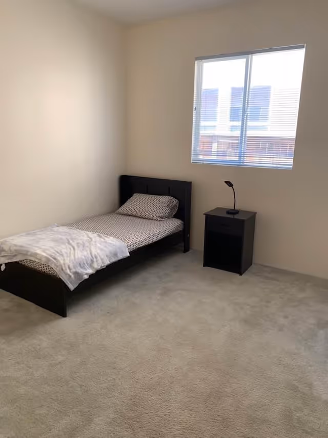 A simple bedroom with a single bed against the wall, featuring a patterned pillow and blanket. Next to the bed is a small black nightstand with a modern black lamp. A window with blinds is on the wall above the nightstand, allowing natural light into the room. The floor is carpeted and the walls are painted a light beige color.