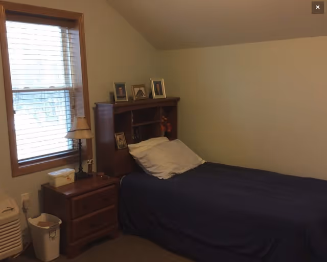 A small bedroom with a single bed covered in a dark blue blanket and a white pillow. The bed has a wooden headboard with shelves holding framed photos and a small flower arrangement. Next to the bed is a wooden nightstand with a lamp, a box, and a trash bin on the floor. A window with blinds is on the left wall.
