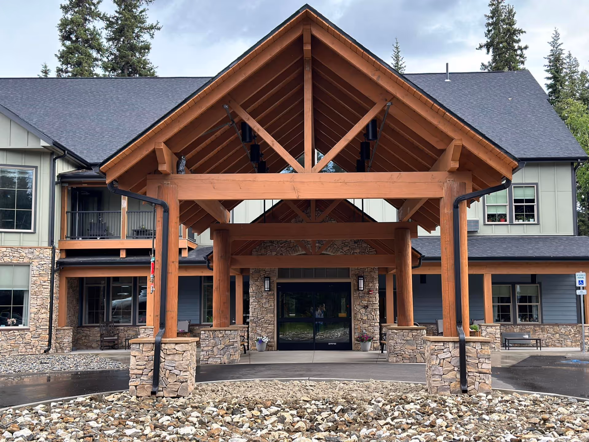 Front exterior view of Timber Creek Senior Living facility featuring a large wooden covered entrance with stone pillars, a paved driveway, and a building with multiple windows and a dark roof surrounded by trees.