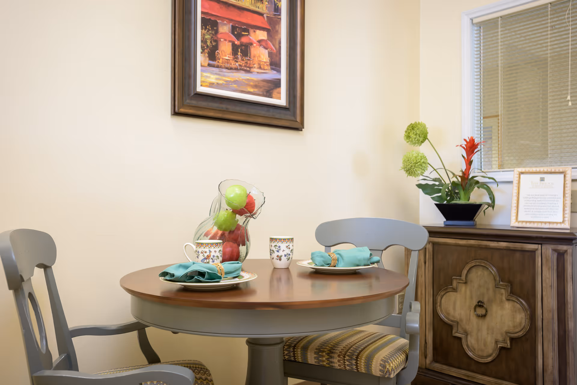 A small round wooden dining table set for two with decorative plates, cups, and teal napkins with napkin rings. A glass pitcher filled with green and red artificial apples is on the table. Two gray wooden chairs with patterned cushions are positioned around the table. In the background, there is a wooden cabinet with a floral arrangement and a framed certificate, and a window with blinds. A framed painting of an outdoor café scene hangs on the beige wall.