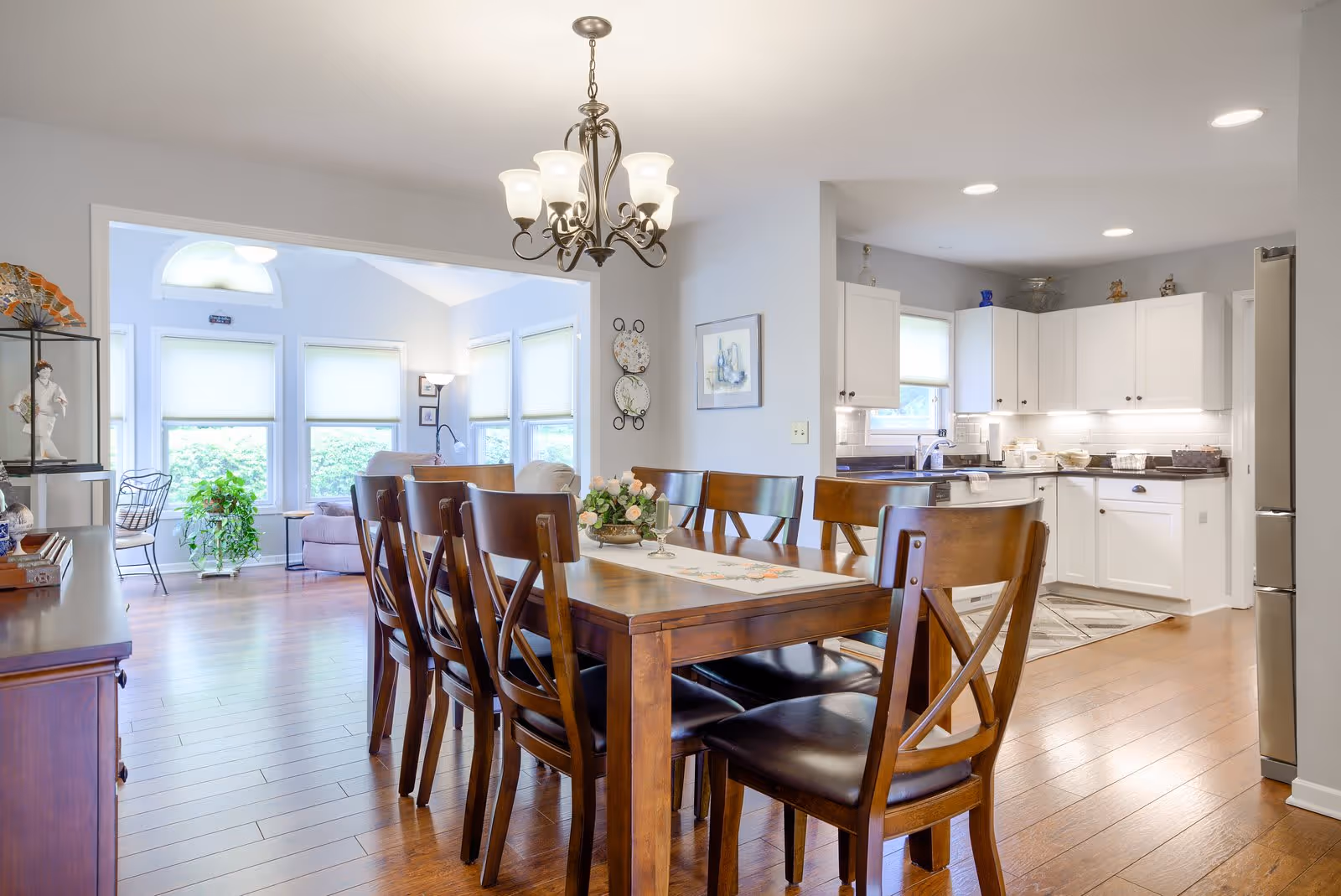 Bright open-plan dining room with a wooden table and chairs leading into a white kitchen and sunlit living area.