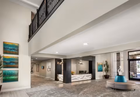 Spacious and modern senior living facility lobby with a reception desk in the center, contemporary artwork on the walls, a round seating area with a decorative sculpture, large windows allowing natural light, and a second-floor balcony overlooking the area.