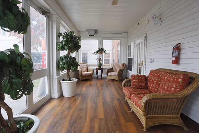 A bright enclosed porch area with wooden flooring, white paneled walls, and large windows. The space is furnished with wicker furniture including a sofa with red patterned cushions, two armchairs, and a small wooden table with a green vase and flowers. There are several potted plants placed around the room, and a fire extinguisher mounted on the wall.