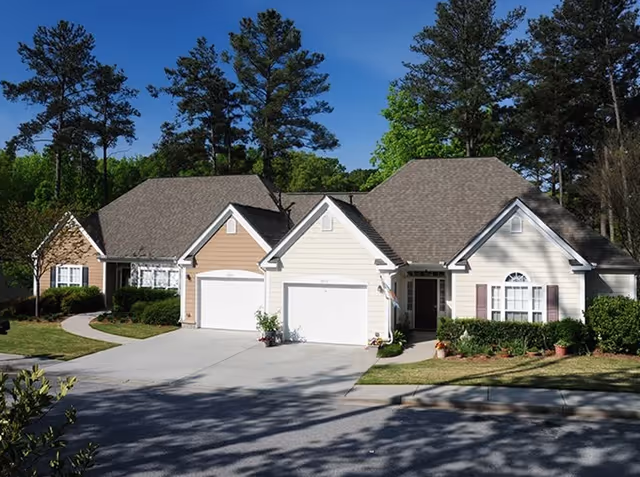 Exterior view of a single-story residential building with two attached garages, surrounded by trees and greenery under a clear blue sky.