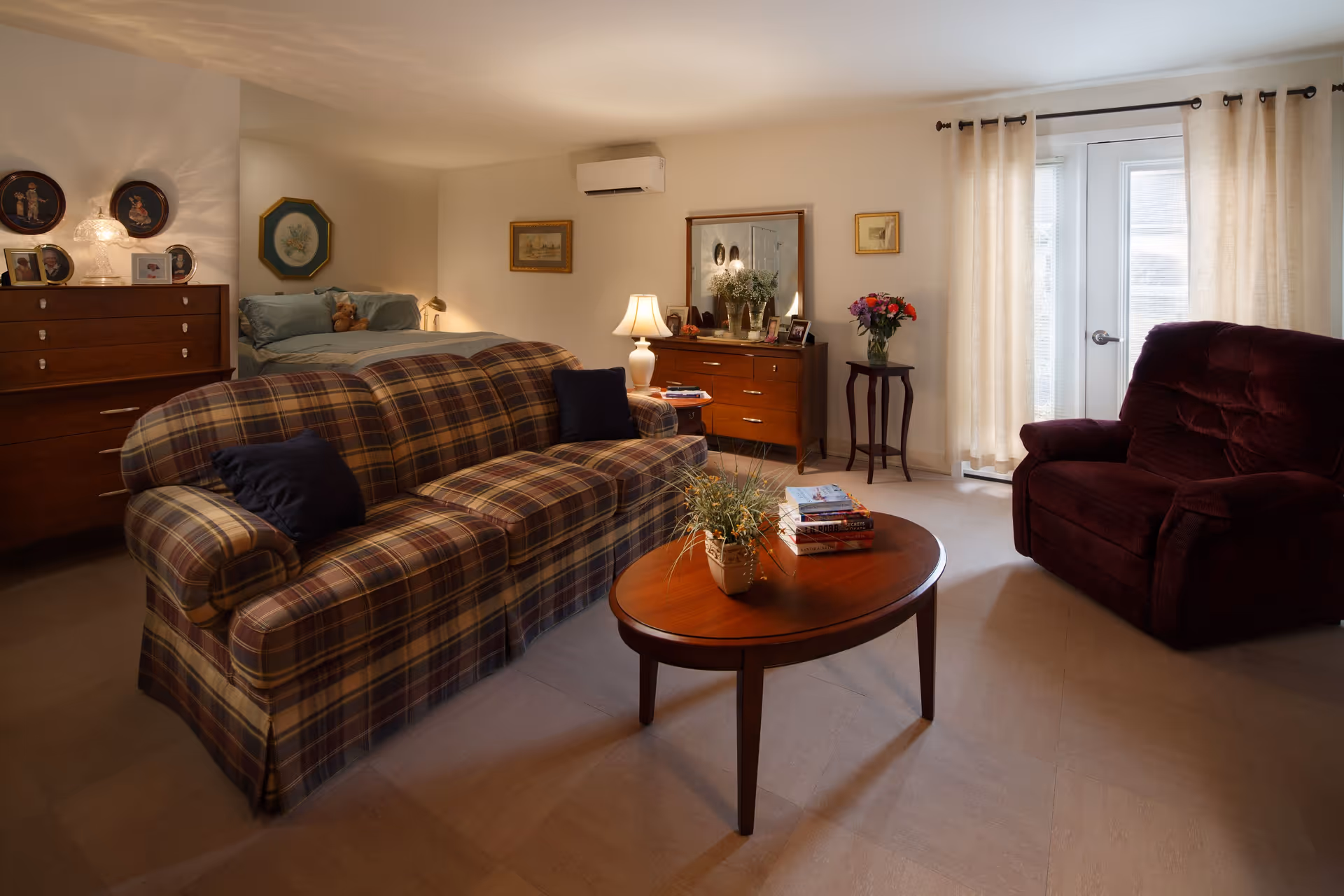 A cozy living area with a plaid sofa, a wooden coffee table with a plant and books, a burgundy armchair, and a dresser with a mirror and lamp. In the background, there is a bed with pillows and framed artwork on the walls. Light filters through sheer curtains covering glass doors.