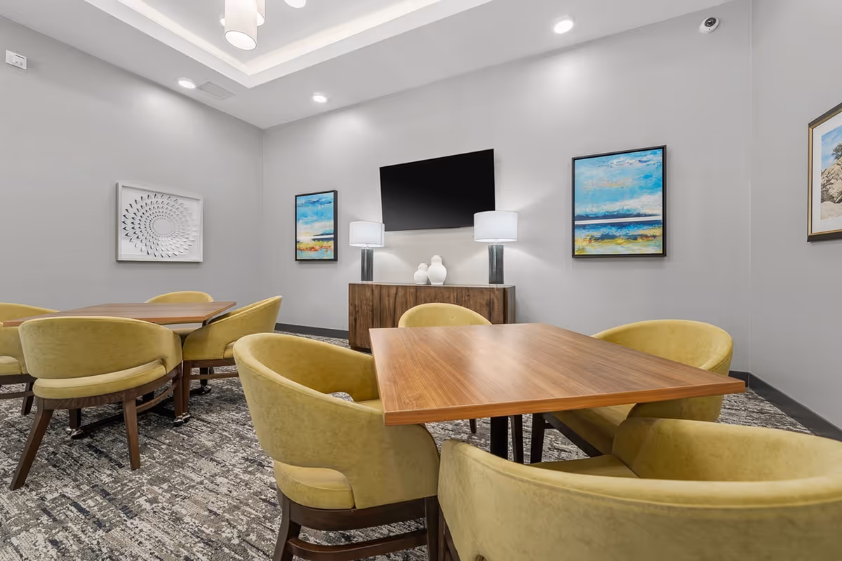 A modern common area with wooden tables and yellow upholstered chairs arranged on a patterned carpet. The walls are light gray and decorated with colorful abstract paintings. A flat-screen TV is mounted on the wall above a wooden cabinet with two table lamps and decorative vases.