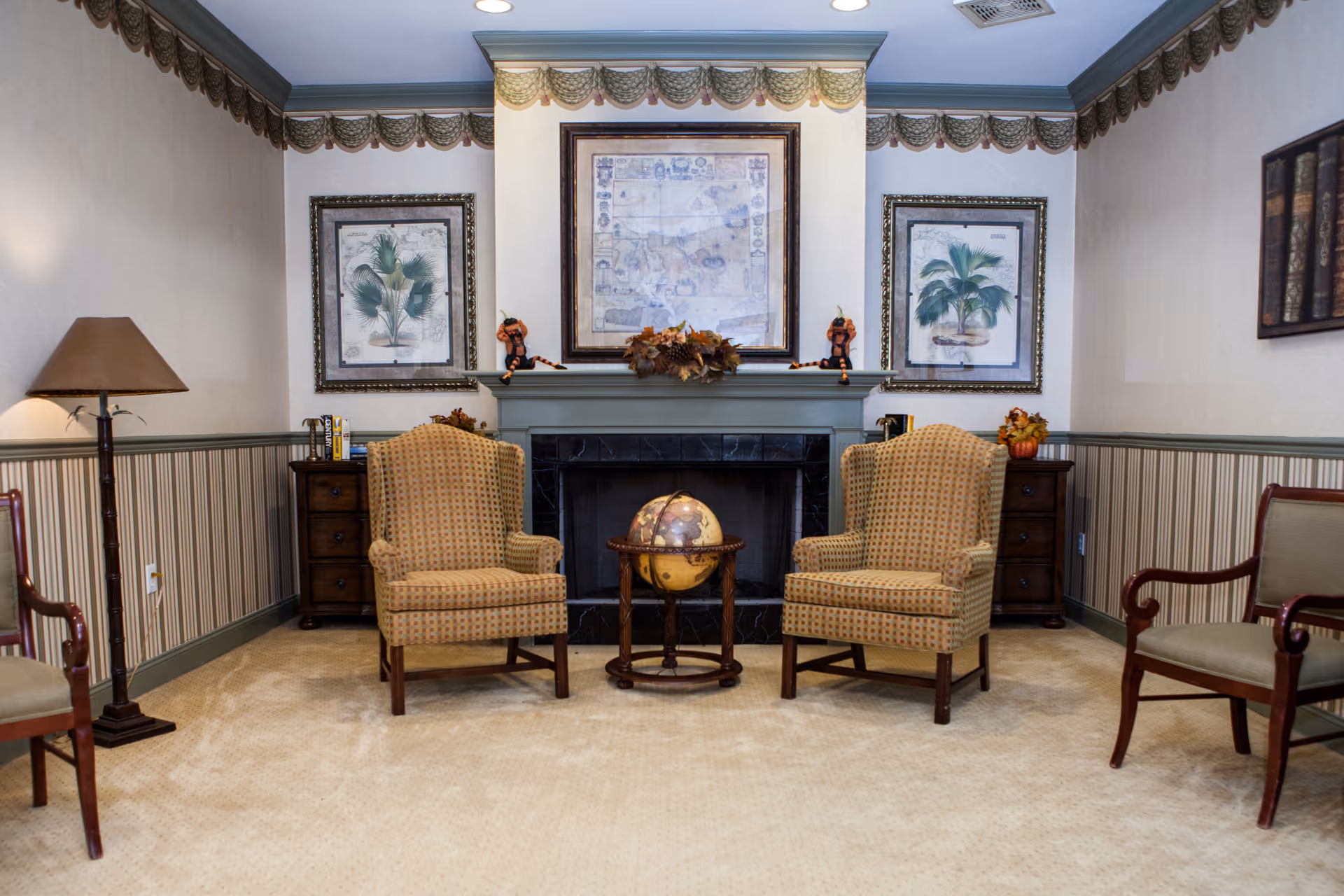 A cozy sitting area with two patterned armchairs facing each other, separated by a small round table holding a globe. The background features a fireplace with a mantel decorated with autumn-themed ornaments and two framed botanical prints on either side. The room has beige carpet, striped wallpaper with a chair rail, and a floor lamp on the left side.