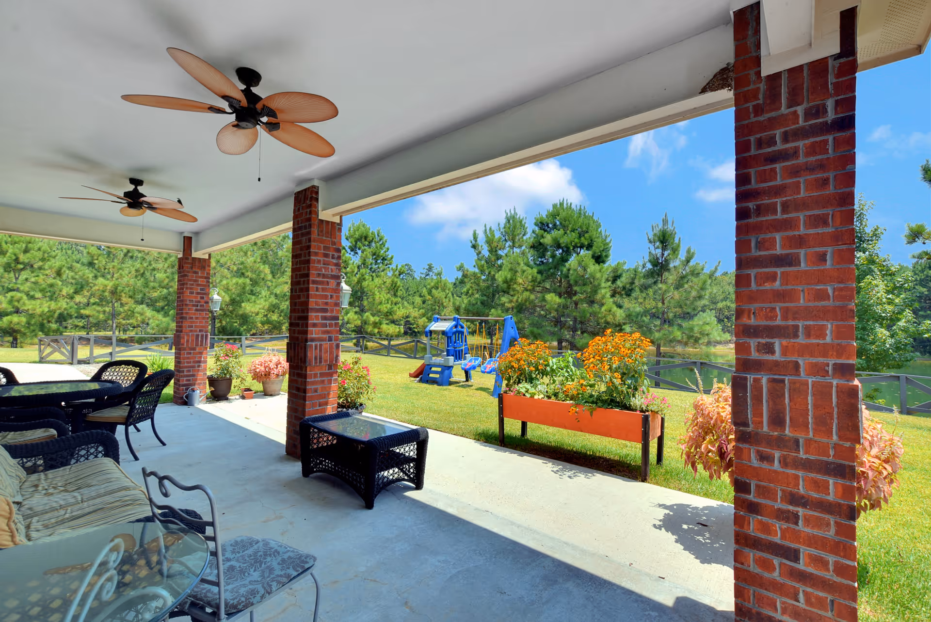 Covered patio with wicker seating, ceiling fans, brick columns, and a view of a grassy yard with planters and a children's playset.