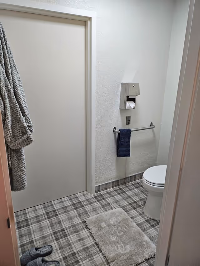 A small bathroom with a closed beige door, a white toilet, a towel rack with a dark blue towel, a wall-mounted toilet paper holder, a gray bathrobe hanging on the door, a pair of gray slippers on the plaid-patterned floor, and a beige bath mat in front of the toilet.