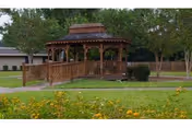 A wooden gazebo with a shingled roof situated in a grassy area with trees and yellow flowers in the foreground. A paved walkway leads up to the gazebo, and there is a building partially visible in the background.