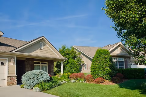 Exterior view of a residential-style building with beige siding and stone accents, surrounded by well-maintained landscaping including bushes, shrubs, and a tree under a clear blue sky.