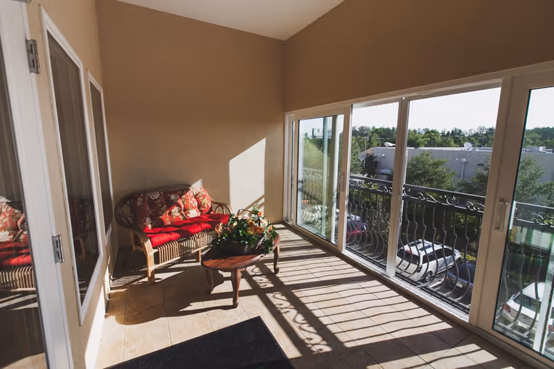 A sunlit enclosed balcony area with large sliding glass doors and windows overlooking a parking lot and trees. The space features a wicker loveseat with red cushions and floral pillows, a small wooden coffee table with a floral arrangement, and tiled flooring.