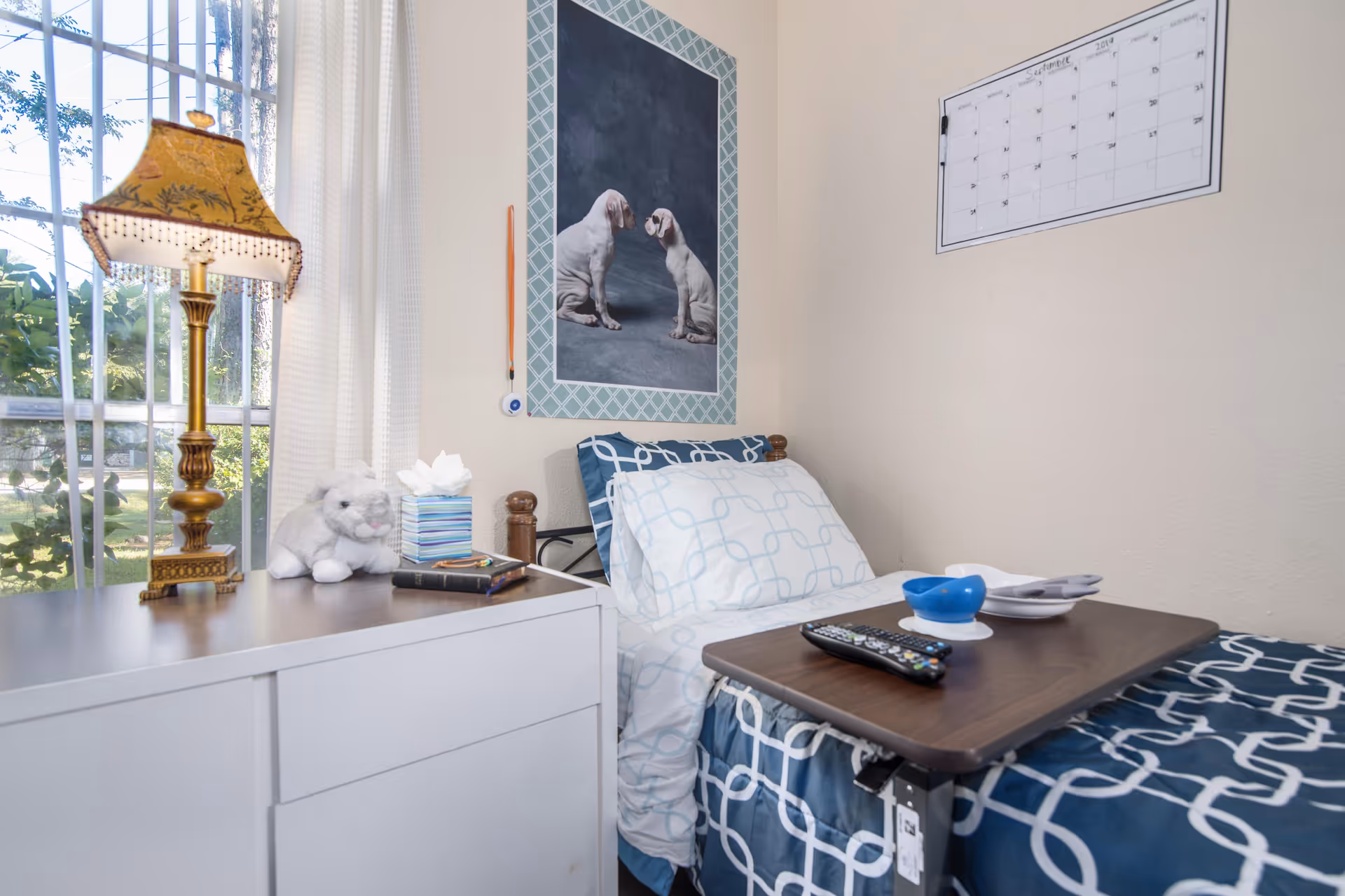 A small bedroom in an assisted living facility with a single bed covered in blue and white patterned bedding. A wooden overbed table holds a remote control, a blue bowl, and a white plate with utensils. Next to the bed is a white dresser with a decorative lamp, a tissue box, a stuffed animal, and a notebook. A window with white curtains lets in natural light. On the wall above the bed is a framed picture of two dogs facing each other and a whiteboard calendar.