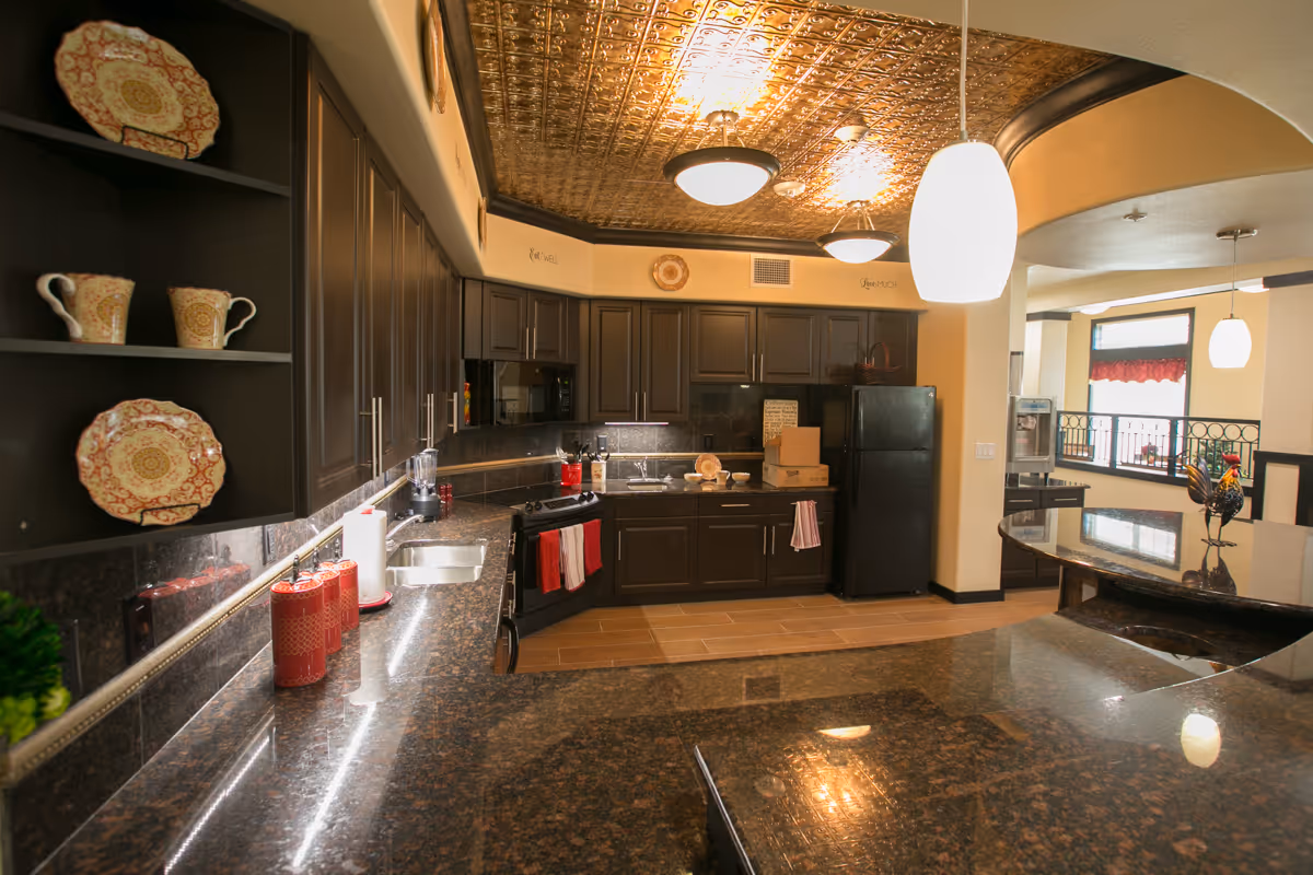 A spacious kitchen with dark wood cabinets, granite countertops, and a decorative tin ceiling. The kitchen features a black refrigerator, stove, microwave, and a double sink. There are decorative plates and mugs on open shelves, red kitchen towels hanging on the oven and cabinet, and pendant lights hanging from the ceiling. In the background, there is a window with a red valance and a countertop with a decorative rooster figurine.