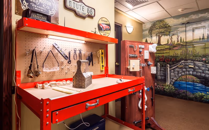 Interior activity room with a red workbench and pegboard tools beside a decorative mural of a stone bridge and landscape.
