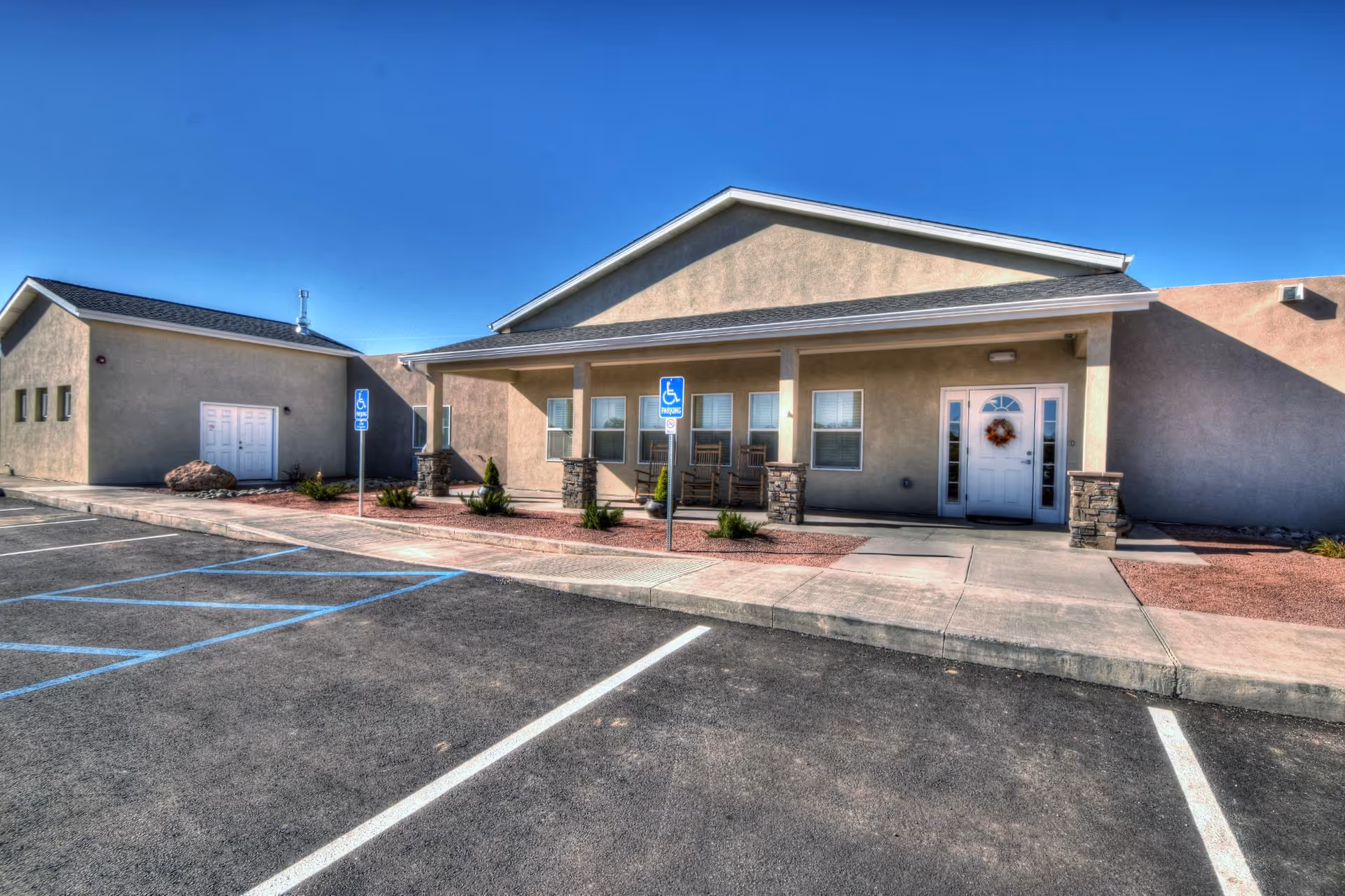 Exterior view of a single-story building with a covered porch featuring rocking chairs. The building has a beige stucco finish and a white door decorated with a wreath. There are two handicap parking signs in front of the building and a clear blue sky overhead.