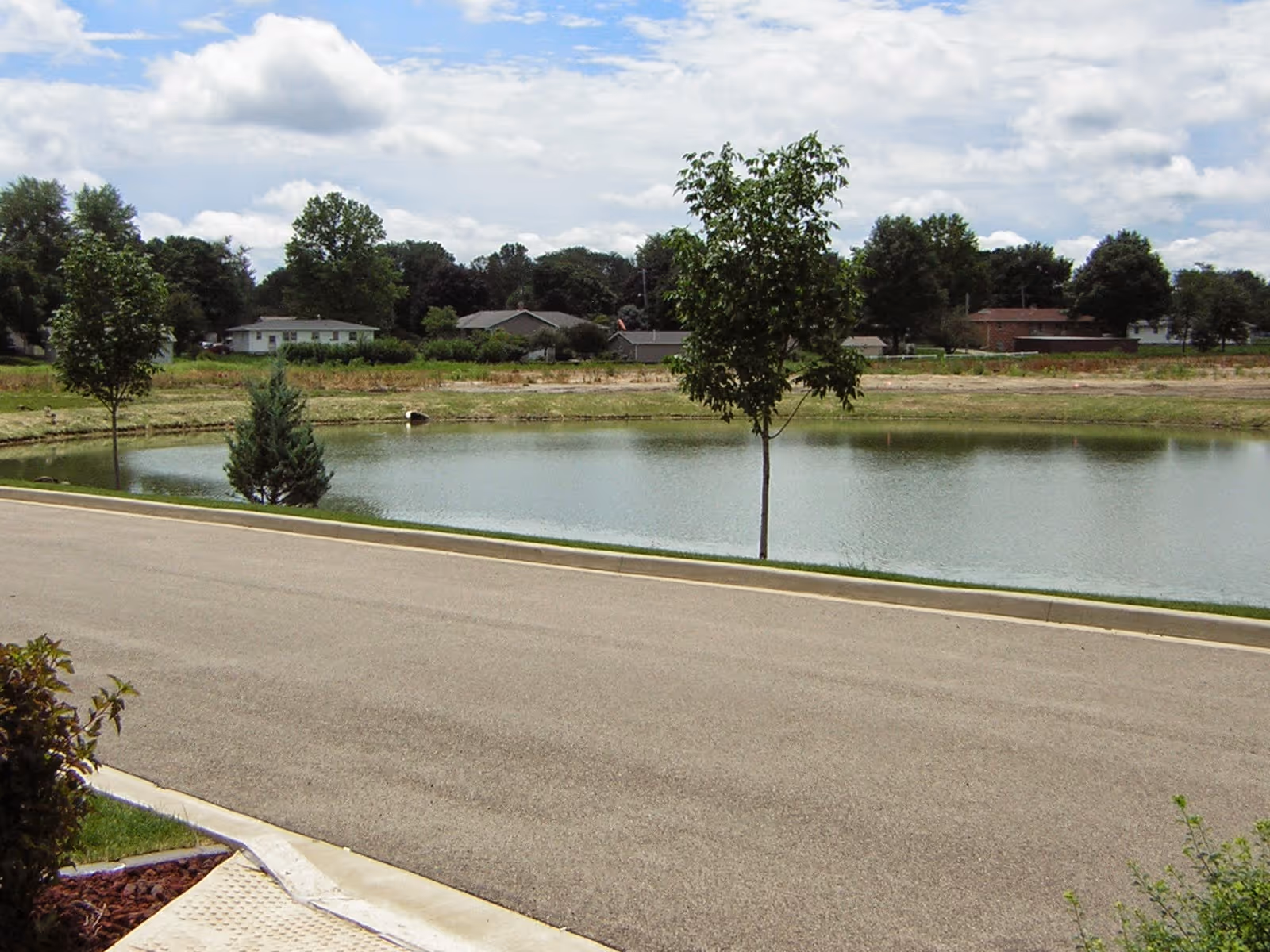 A paved road runs alongside a small pond with young trees and houses in the background under a partly cloudy sky.