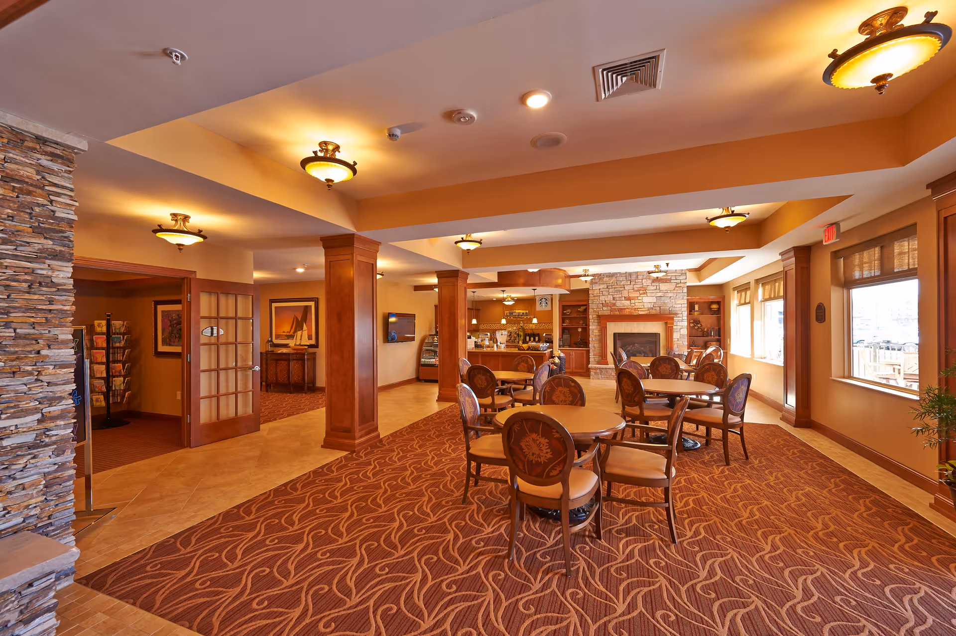 A warm and inviting common area in a senior living facility featuring round tables with chairs, a stone fireplace, wooden columns, and soft lighting. The room has large windows allowing natural light to enter and a carpet with a wavy pattern covering part of the floor.