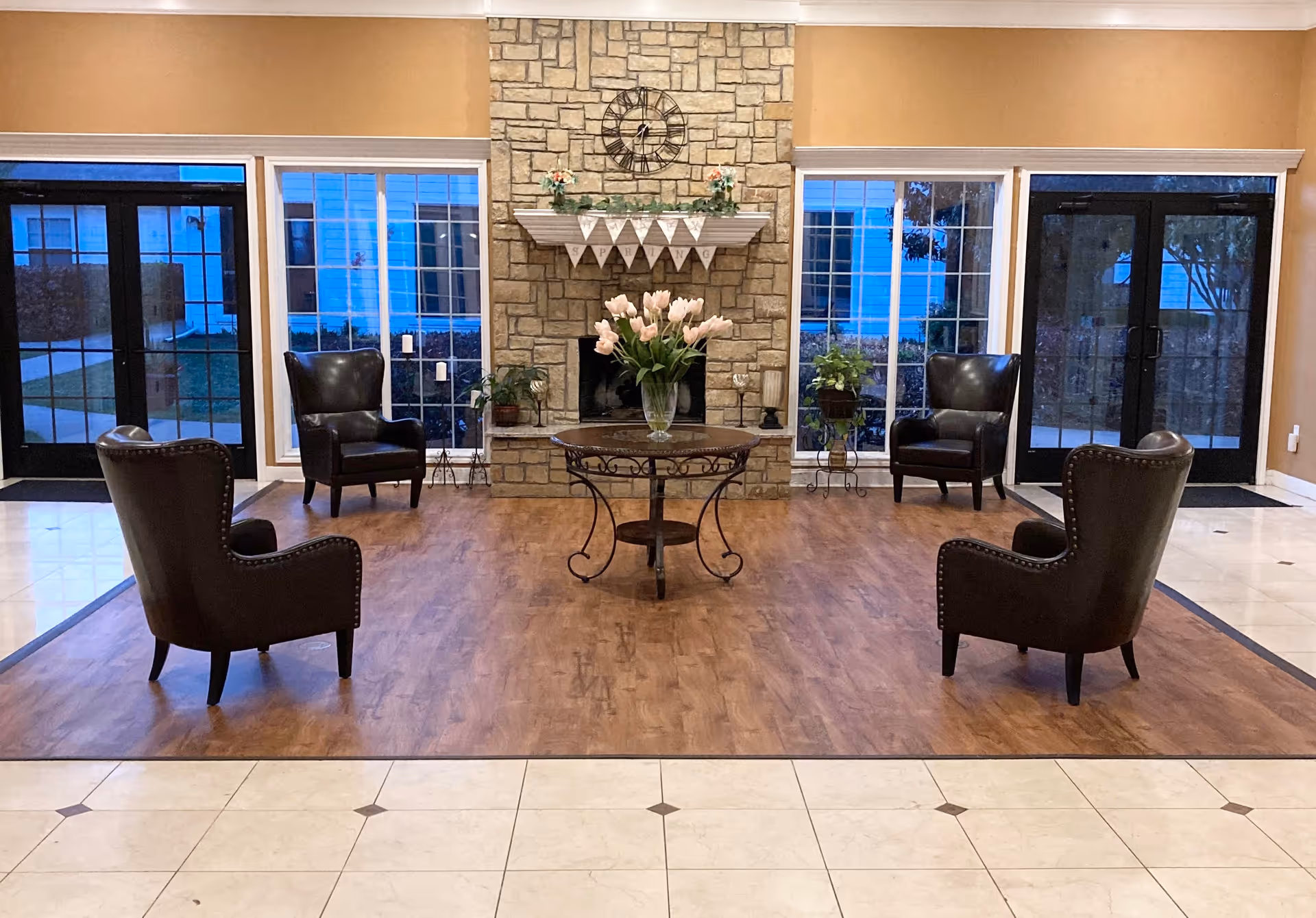 A cozy seating area in an assisted living facility with four dark leather armchairs arranged around a round wooden table holding a vase of pink tulips. The background features a stone fireplace with a clock above it and a banner hanging from the mantel. Large windows and glass doors on either side provide a view of the outside.