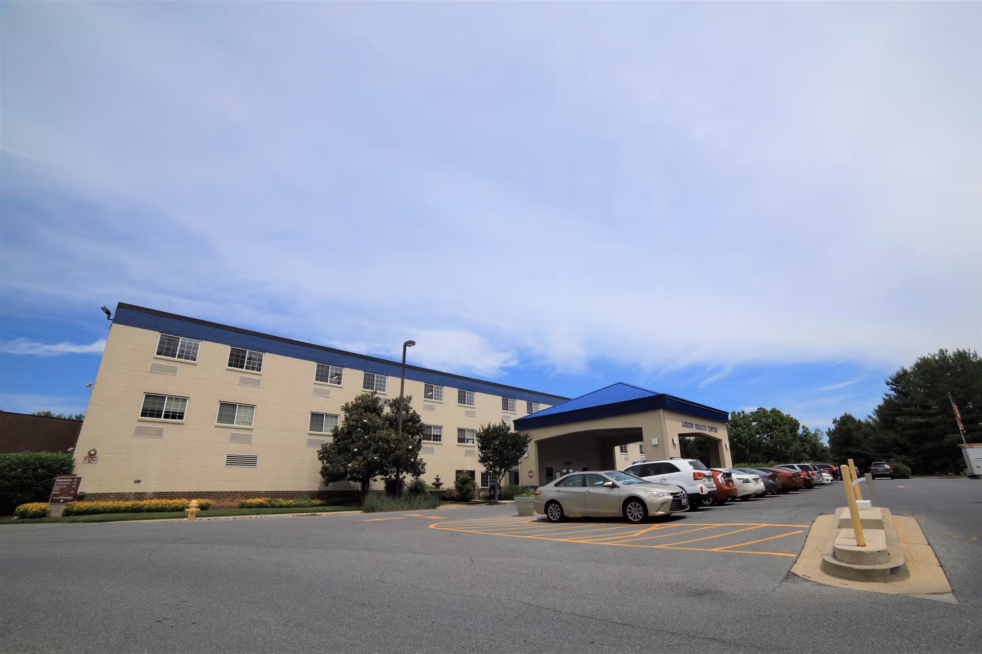 Front exterior of a three-story beige senior living building with a blue roof and covered entrance, a parking lot with cars and trees in front.
