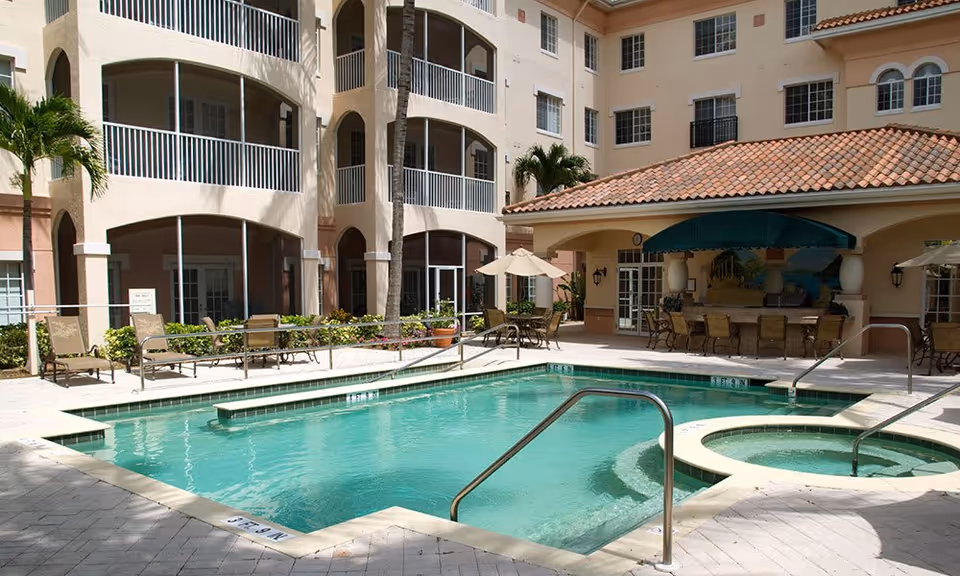 Outdoor swimming pool area at Terracina Senior Living with lounge chairs, tables with umbrellas, and a covered patio with seating. The pool is surrounded by a multi-story building with balconies and palm trees.