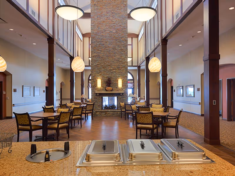 Spacious dining area with multiple tables and chairs arranged around a central stone fireplace. The room features high ceilings with large hanging light fixtures and tall windows allowing natural light. In the foreground, there is a countertop with three covered food warmers and a sink.