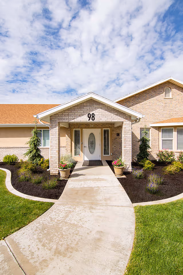 Front entrance of a single-story brick building with the number 98 above the door. The entrance has a covered porch with two potted plants on either side of the walkway. The surrounding landscaping includes green grass, small bushes, and trees under a partly cloudy sky.