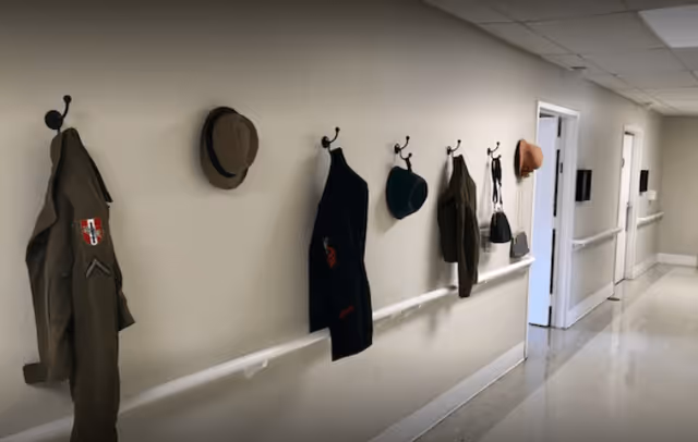 A hallway with a light-colored wall featuring several hooks holding various hats, jackets, and bags. The floor is shiny and light-colored, and there are doors and handrails along the hallway.