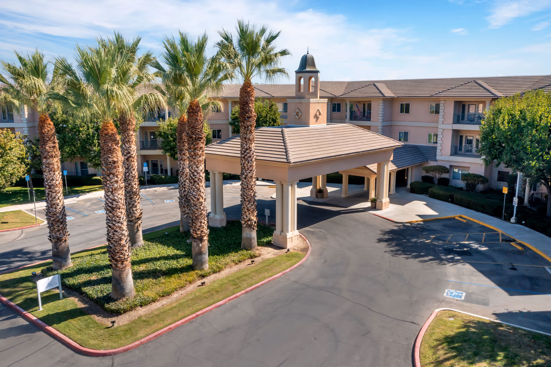 Exterior view of Brookdale Riverwalk senior living facility showing a covered entrance with columns, surrounded by palm trees and a parking area with marked handicap spaces under a clear blue sky.