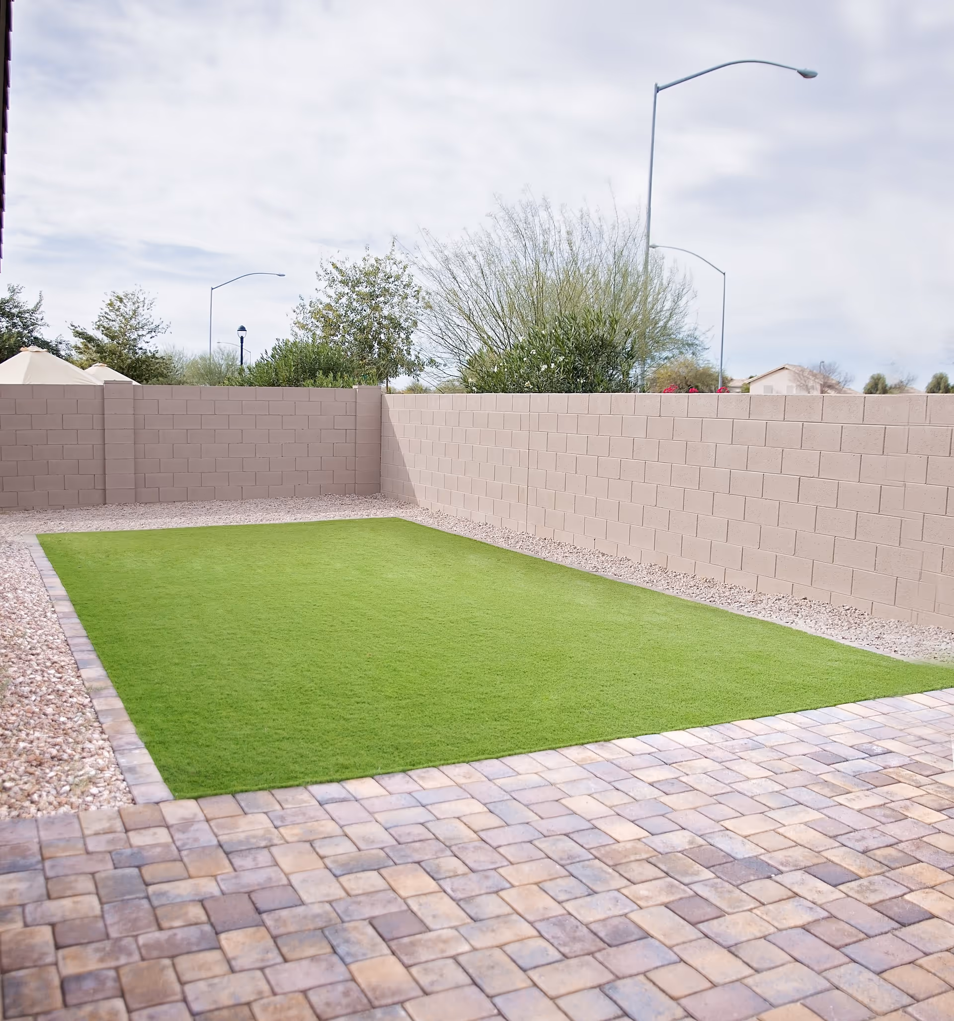 A small outdoor patio area with a paved stone floor surrounding a rectangular patch of green artificial grass, enclosed by a beige brick wall. Trees and street lamps are visible beyond the wall under a cloudy sky.