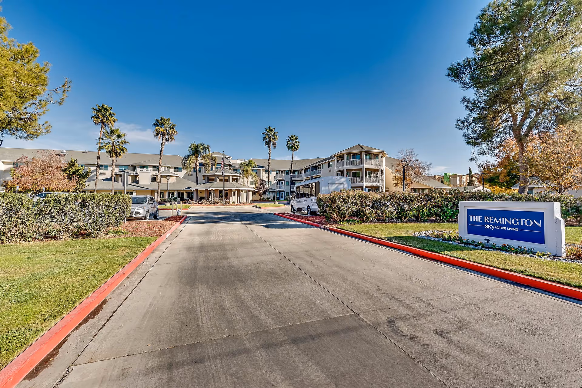 Front exterior view of The Remington Senior Living facility with a driveway leading to the entrance, surrounded by palm trees, bushes, and a clear blue sky.