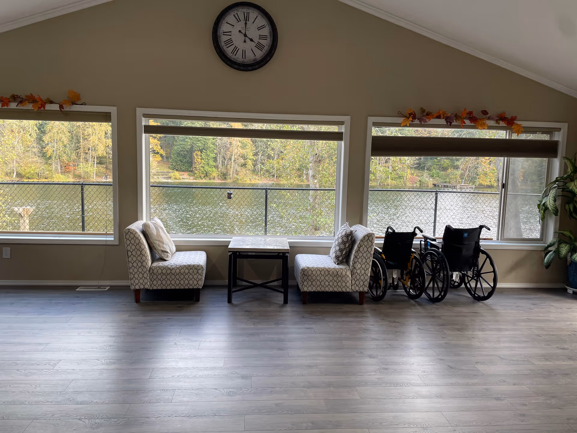 A bright room with three large windows showing a view of a lake and trees outside. Inside, there are two patterned chairs with pillows facing each other across a small table. Two wheelchairs are positioned near the right window. A clock hangs on the wall above the middle window, and autumn leaf decorations adorn the window frames. A potted plant is visible on the far right side of the room.