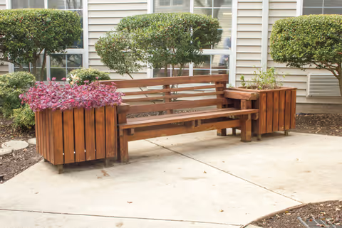 A wooden bench with built-in planters on both ends, containing purple and green plants, situated on a concrete patio in front of a building with beige siding and windows. There are neatly trimmed bushes behind the bench.