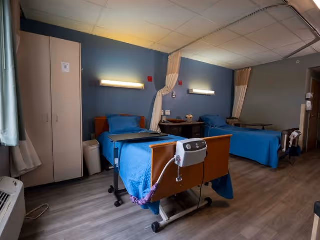 A shared patient room in a rehabilitation facility with two hospital beds covered in blue blankets. Each bed has a bedside table and medical equipment. The room has blue walls, overhead lighting, a wardrobe, and a curtain divider between the beds.