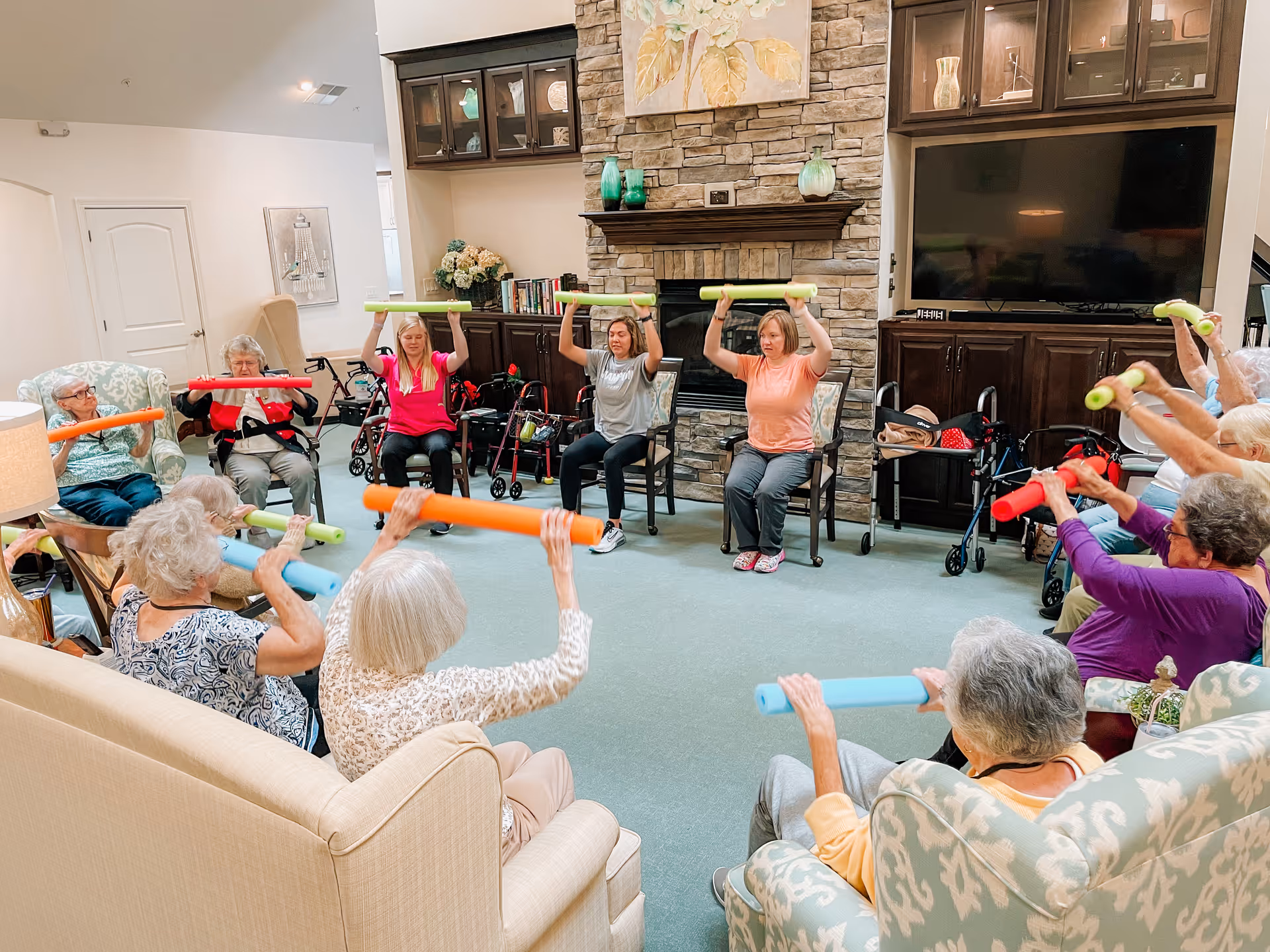 Residents and staff seated in a common living room circle lifting colorful foam noodles during a group exercise session.