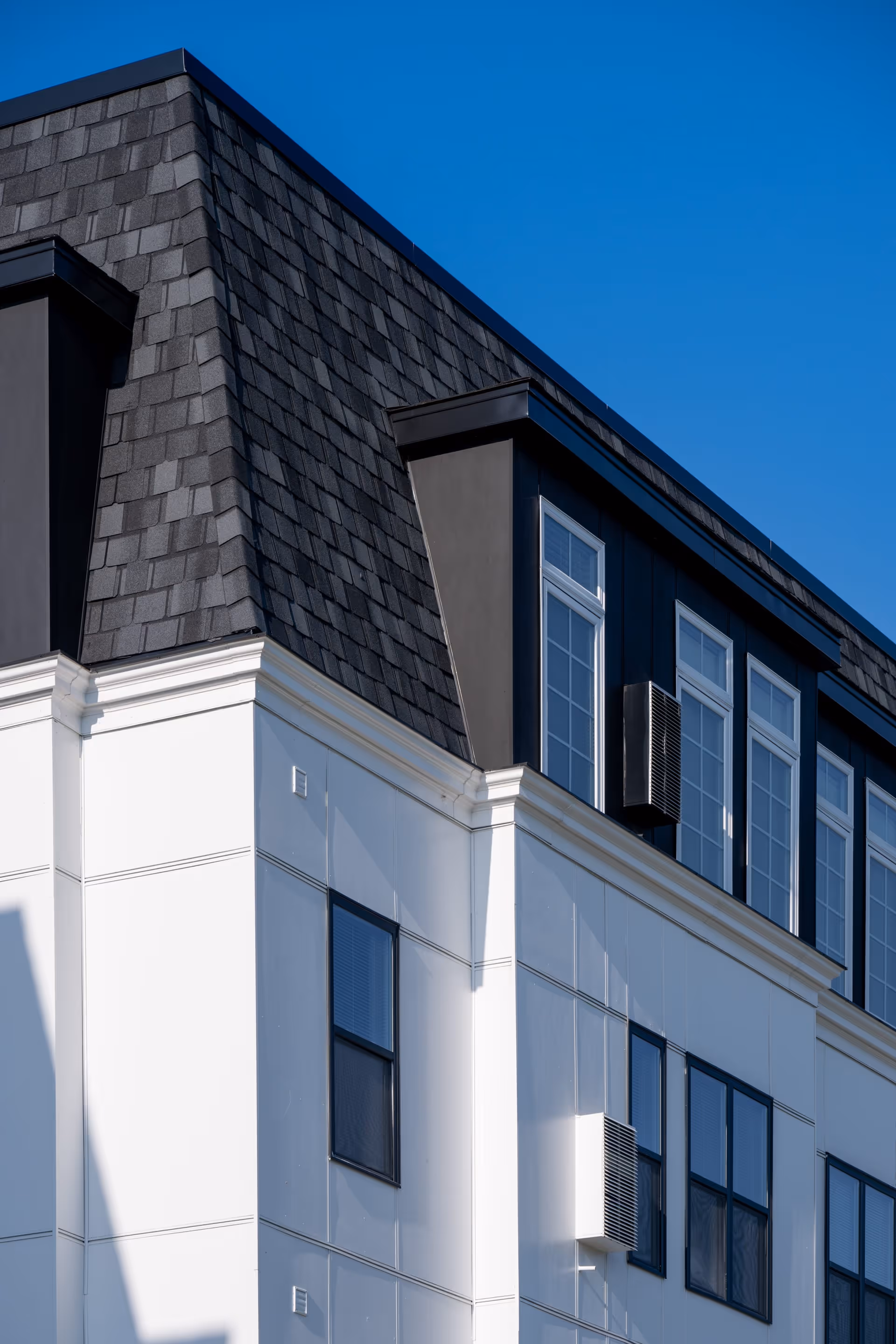 Close-up view of the upper corner of a modern building with white walls, black framed windows, and a dark shingled mansard roof under a clear blue sky.