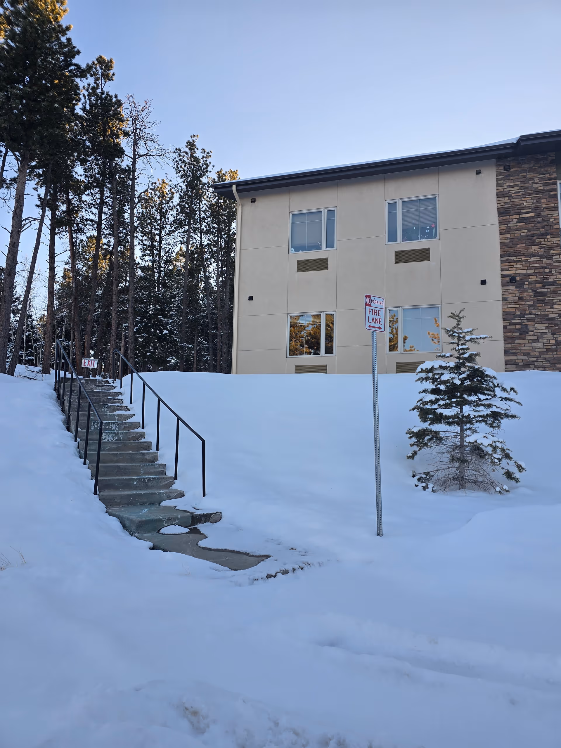 Snow-covered outdoor area with a concrete staircase leading up to a building. The building has beige walls with stone accents and several windows. There is a small pine tree covered in snow near a 'No Parking Fire Lane' sign. Tall pine trees are visible in the background under a clear blue sky.