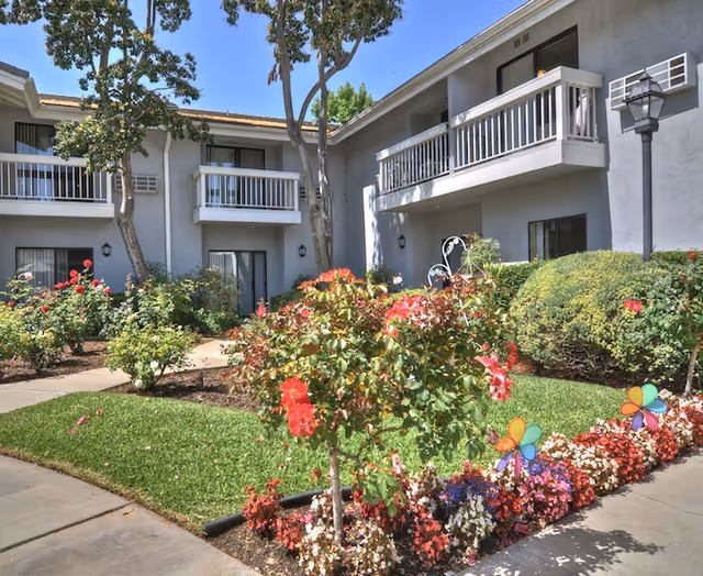 Outdoor courtyard area of a senior living facility with well-maintained flower beds, green grass, and small trees. The building has two stories with balconies and windows overlooking the garden. Colorful pinwheel decorations are placed among the flowers.