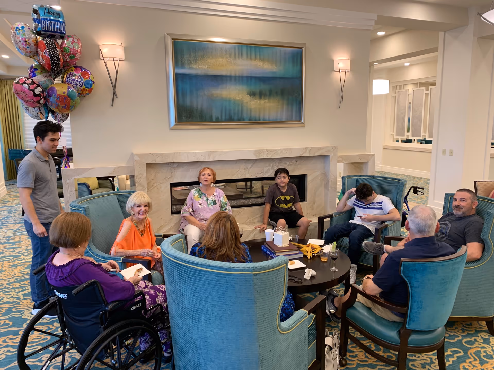 A group of people, including elderly individuals and younger adults, gathered in a comfortable living room area with teal armchairs arranged around a circular coffee table. There is a fireplace with a large abstract painting above it, and a cluster of colorful birthday balloons in the background. The setting appears warm and inviting, with soft lighting and carpeted floors.